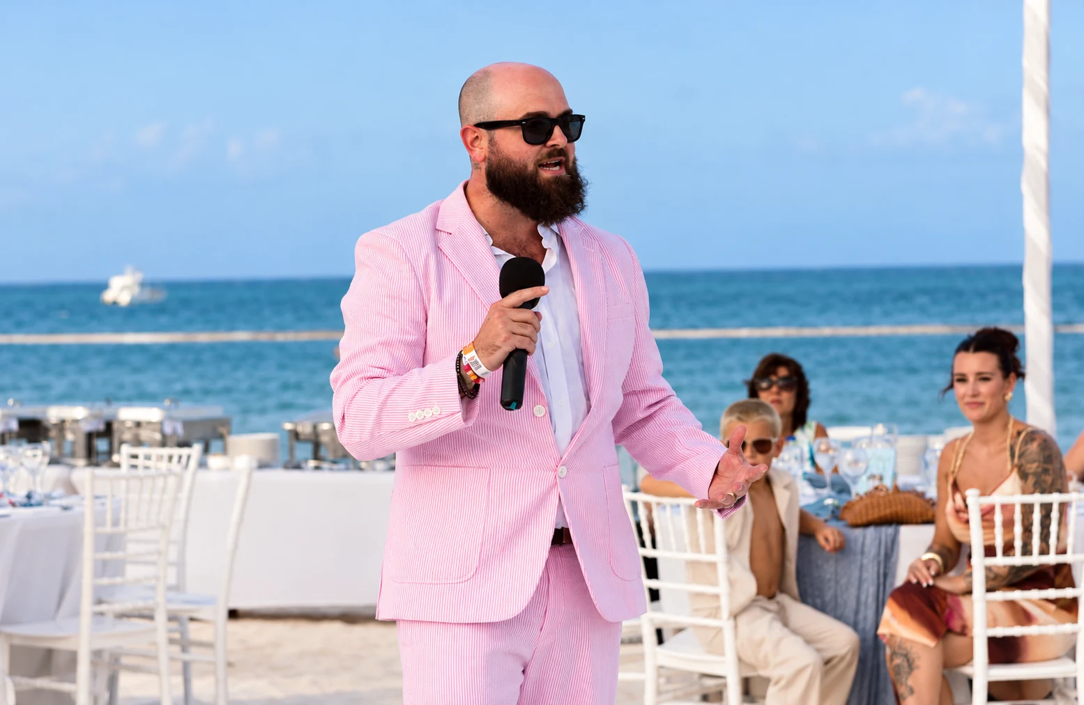 Kyle in a pink suit holding a microphone at a beach wedding reception